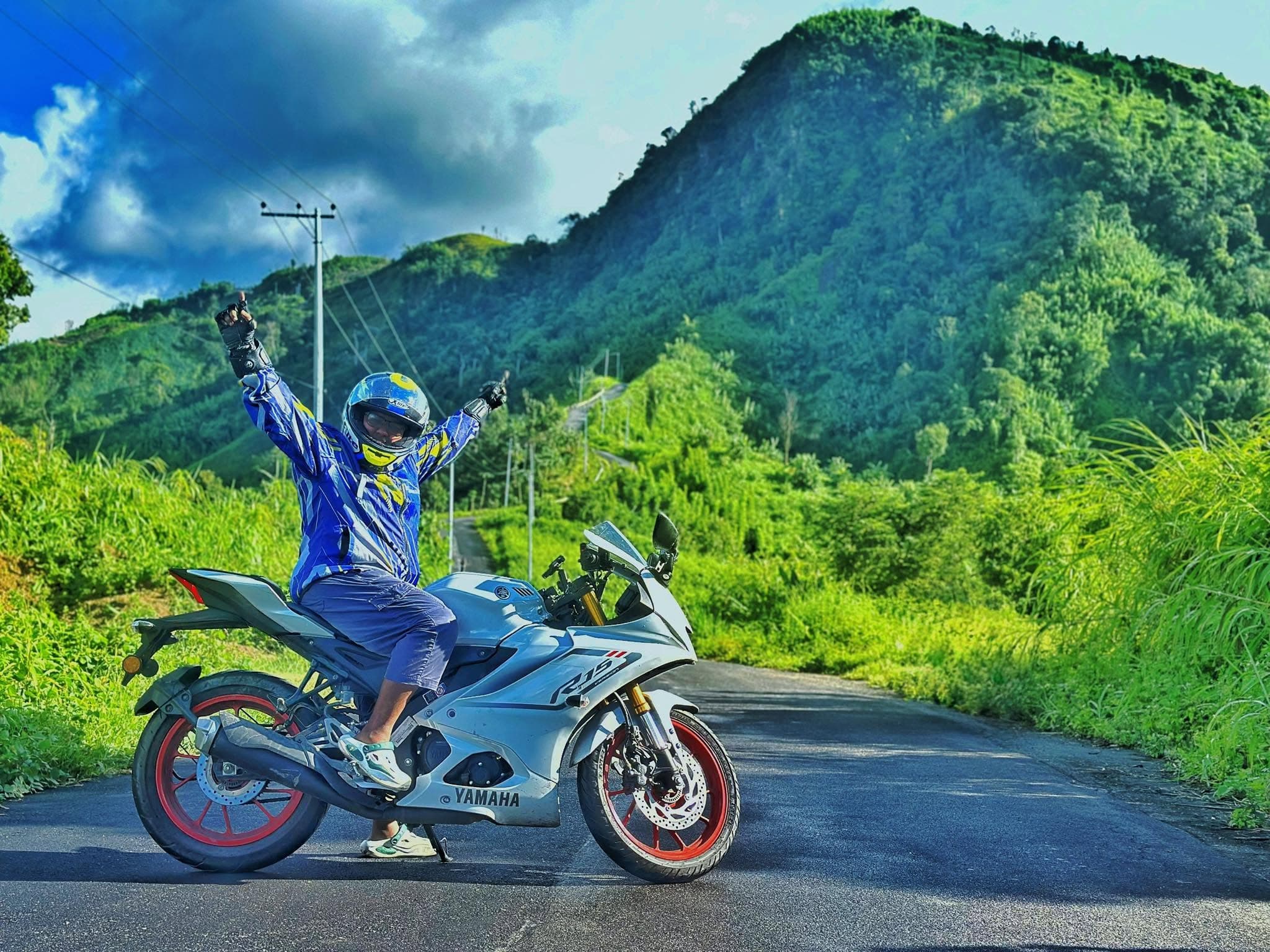 Lina on a Yamaha R15 with her arms raised, riding a green hill road. Landscape.